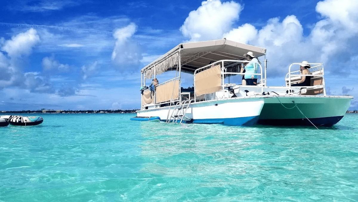 A white and blue catamaran with a tan canopy and fringe trim floats on crystal-clear turquoise water under a bright, cloudy sky.