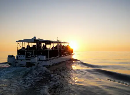 A boat filled with passengers travels across calm water toward a bright, glowing sun on the horizon.