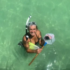 A smiling woman wearing snorkeling gear standing in clear water, holding up a small net with a sea urchin inside.
