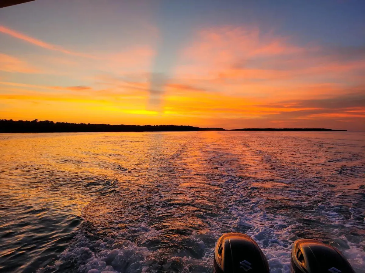 A view from the back of a moving boat showing a white wake trailing toward a vibrant orange and yellow sunset over a dark shoreline.