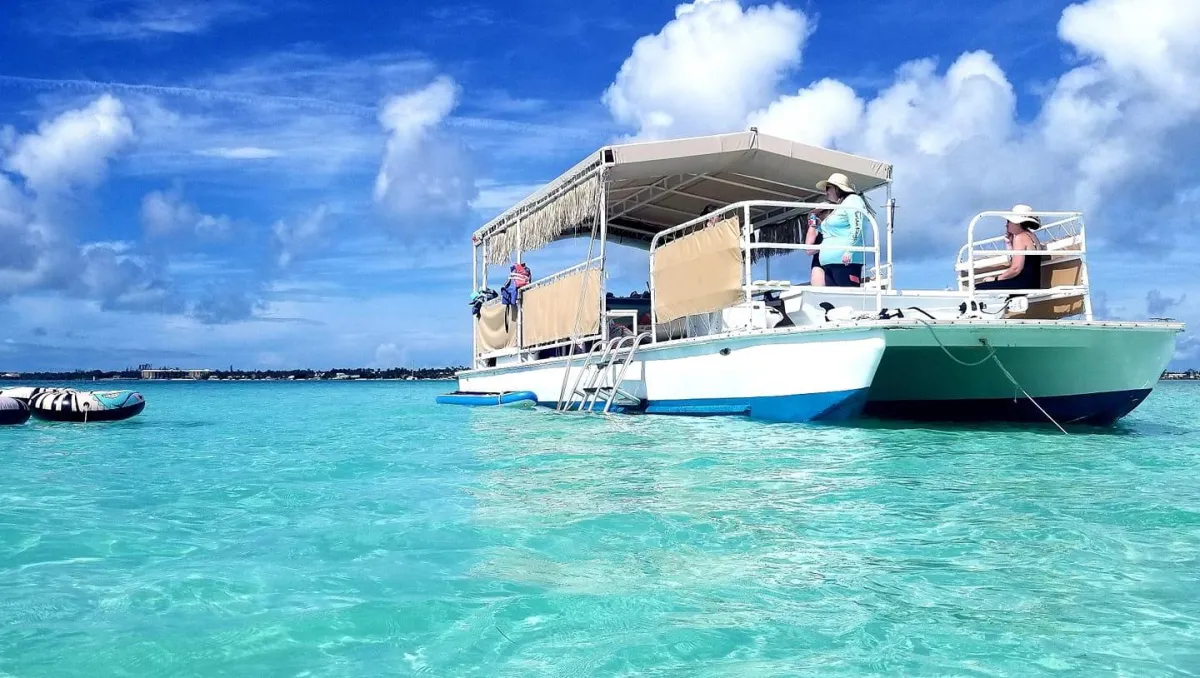 A white and blue catamaran with a tan canopy and fringe trim floats on crystal-clear turquoise water under a bright, cloudy sky.