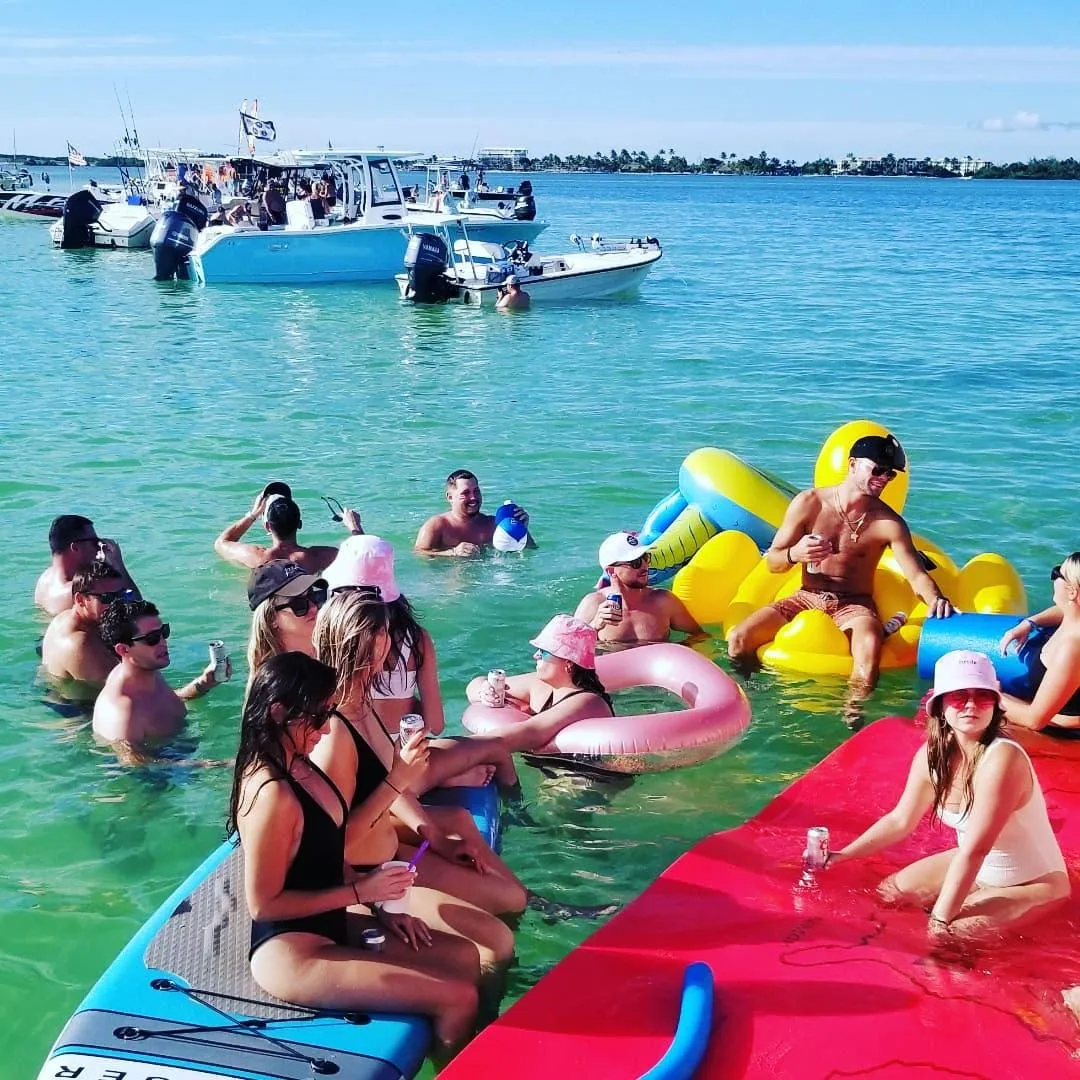 A group of people relax and socialise in shallow turquoise water with drinks, paddleboards, and colourful floats.
