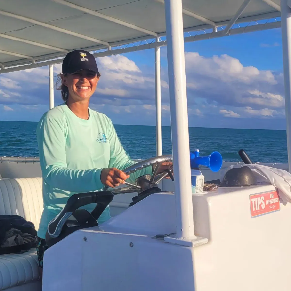 A smiling woman in a teal long-sleeve shirt and baseball cap steers a boat on a calm blue ocean under a cloudy sky.